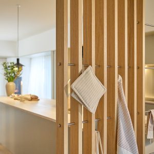 Divisória ripada em carvalho da WoodUpp com barramentos metálicos encaixáveis formando ganchos, utilizada como suporte para luvas de panela e panos de cozinha; ao fundo, bancada clara e objetos decorativos, mostrando aplicação prática e estética do sistema modular WoodUpp na cozinha contemporânea.
