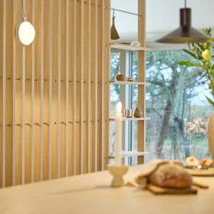 Painel ripado de carvalho WoodUpp com pegas metálicas e prateleiras modulares encaixadas, criando divisória e estante junto à janela. Pequenos objetos decorativos em madeira, uma escova pendurada e um candeeiro circular na parede; em primeiro plano, vela acesa e pão sobre a mesa.