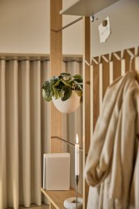 Painel modular WoodUpp em madeira clara estilo carvalho com lâminas verticais, prateleira inferior, haste metálica e ganchos. Vaso branco semi‑esférico suspenso no suporte, livros empilhados, castiçal fino com vela acesa sobre apoio de pedra clara e casaco pendurado, tudo em ambiente neutro com cortinas.