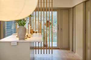 Interior de cozinha minimalista com bancada e vaso; destaque para um painel divisório de ripas em madeira da WoodUpp, equipado com barra e ganchos WoodUpp para pendurar toalhas e sacola de rede, frente a uma porta de correr envidraçada e chão de madeira claro.