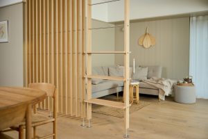 Divisória WoodUpp em ripas de madeira clara (carvalho) com pés metálicos, integrando prateleiras finas e hastes de metal, separando sala e sala de jantar. Ambiente neutro com sofá cinza, mesa lateral em rattan, manta e detalhes decorativos visíveis ao fundo.