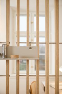 Divisória de ripas verticais em madeira clara da WoodUpp, com prateleira modular cinza fixada e pequenos acessórios: um vaso cilíndrico metálico e uma tigela de madeira. Ambiente claro com janela ao fundo, cadeira e mesa parcialmente visíveis, estilo minimalista e funcional.