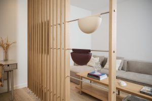 Divisória de ripas de madeira WoodUpp separa ambiente, com barras metálicas e dois cachepôs suspensos WoodUpp em forma de meia‑lua — um creme e outro bordô. Ao fundo, sofá cinza, mesa de centro com livros e vaso de vidro, e mesa lateral com arranjo seco.