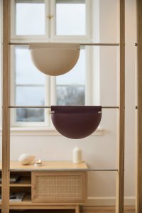 Imagem mostra elementos WoodUpp: dois vasos suspensos em formato meia lua, um bege claro e outro vinho escuro, encaixados em varões de metal dentro de uma estrutura de madeira. Ao fundo há um móvel baixo, vela e janela, criando ambiente interior minimalista e acolhedor.