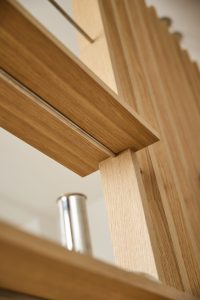 Close-up do produto WoodUpp: painéis ripados e prateleira em carvalho claro com acabamento natural, fixados por perfil cilíndrico metálico e suporte tubular cromado. Detalhe realça veios da madeira, união entre ripas verticais e prateleira horizontal, design elegante e modular para divisórias.