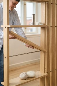 Imagem mostra um homem ajustando uma estante modular WoodUpp em carvalho com ripas verticais, prateleiras ajustáveis e um painel basculante apoiado por varões metálicos; na prateleira inferior estão novelos de lã com agulha e na superior um copo metálico e um sabonete.