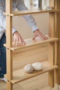 Prateleira modular WoodUpp em carvalho natural com estrutura vertical, prateleiras encaixáveis, parafusos aparentes e acabamento aveludado. Mãos ajustam uma das tábuas enquanto no nível inferior repousam novelos de lã claros e agulhas de tricô; objetos decorativos (pedra e acessório metálico) no topo.