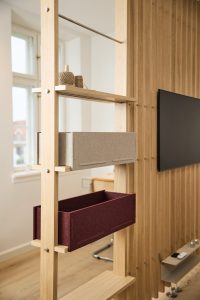 Divisória modular WoodUpp em madeira clara com prateleiras e trilhos, exibindo duas caixas organizadoras de feltro WoodUpp — uma cinza e outra bordô — encaixadas nas ripas. No topo há rolos de corda, ao fundo painel ripado e televisão montada na parede.