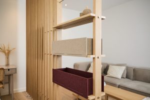 Divisória modular de madeira WoodUpp com prateleiras e duas caixas organizadoras de feltro WoodUpp — uma bege e outra vinho — encaixadas nas prateleiras. Estrutura em madeira clara com parafusos visíveis, objeto decorativo no topo; ao fundo sofá e área de trabalho.