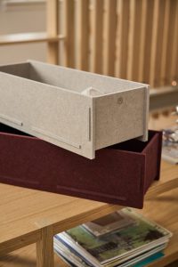 Imagem WoodUpp mostra duas gavetas organizadoras em feltro modular: uma bege clara, suspensa, com logotipo gravado, e outra bordô apoiada numa mesa de madeira. Design encaixável com abas laterais, interior amplo para roupas ou acessórios, acabamento suave e estética minimalista.