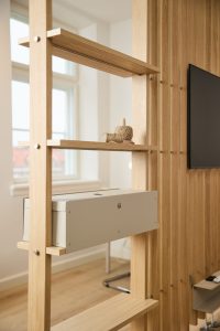 Divisória modular em madeira clara da WoodUpp com prateleiras horizontais e painel ripado; destaque para a caixa metálica organizadora de cabos WoodUpp embutida nas prateleiras, detalhes em parafusos aparentes e acabamento em carvalho, em ambiente iluminado com televisão ao fundo.