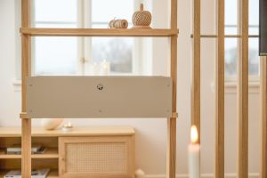 Divisor modular WoodUpp em ripas de carvalho claro, com prateleira superior exibindo rolos de corda, e inserto metálico bege WoodUpp fixado entre as ripas (painel retangular com parafusos e saída de ventilação). Cena com velas acesas e móvel rústico ao fundo.
