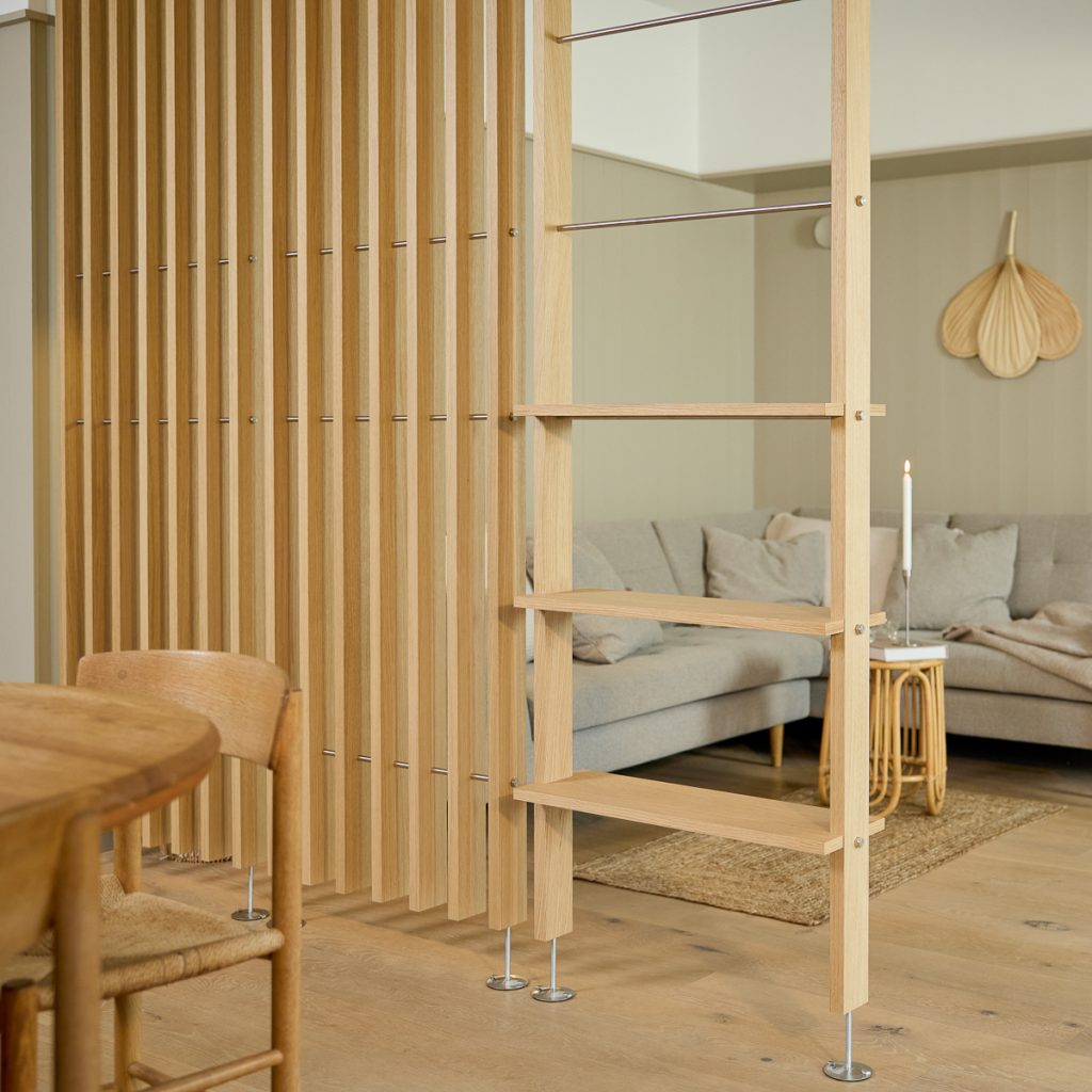 Divisor de ambientes ripado em madeira clara da WoodUpp com pés metálicos, acompanhado de uma estante/escada com prateleiras horizontais WoodUpp; cena de sala de estar com sofá cinza, mesa lateral em rattan e vela acesa, transmitindo estilo escandinavo e luz natural.
