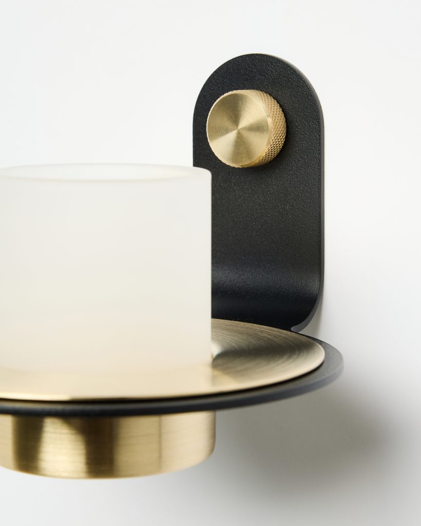 Close-up de arandela WoodUpp: vidro leitoso cilíndrico sobre prato e base em latão escovado dourado, suporte de parede em metal preto fosco com topo arredondado e botão moleteado de latão. Design minimalista que destaca texturas metálicas e acabamento moderno e elegante.
