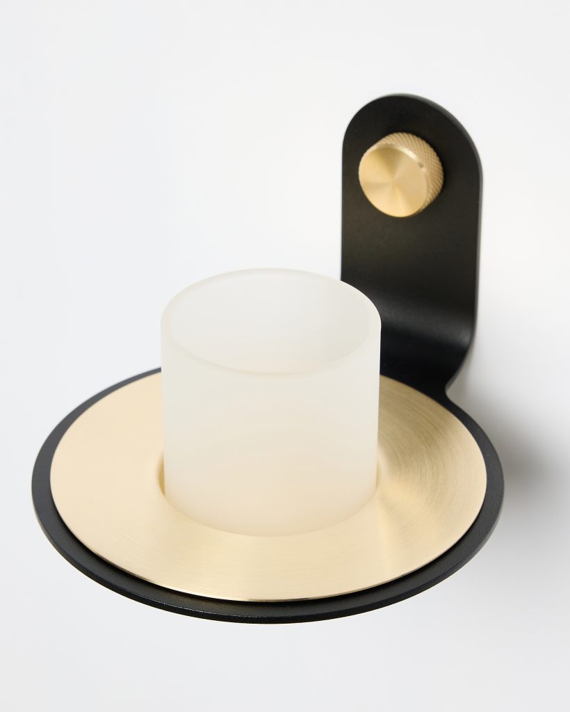 Suporte de vela de parede WoodUpp em metal preto, com prato circular dourado escovado e copo para vela em vidro fosco translúcido; detalhe de ajuste em botão metálico dourado texturizado. Com acabamento premium e linhas contemporâneas, fotografado sobre fundo branco.