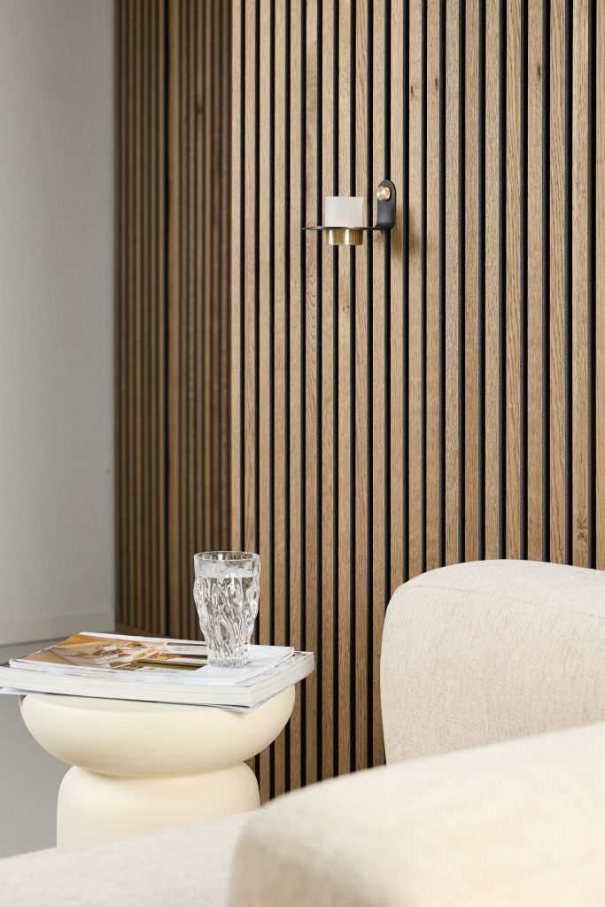 Painel ripado WoodUpp em madeira clara, com lâminas verticais e vãos escuros formando textura linear, instalado atrás de um sofá bege. Detalhe de castiçal de parede preto e latão com vela, mesa lateral creme com revista e copo de água, ambiente minimalista e acolhedor.