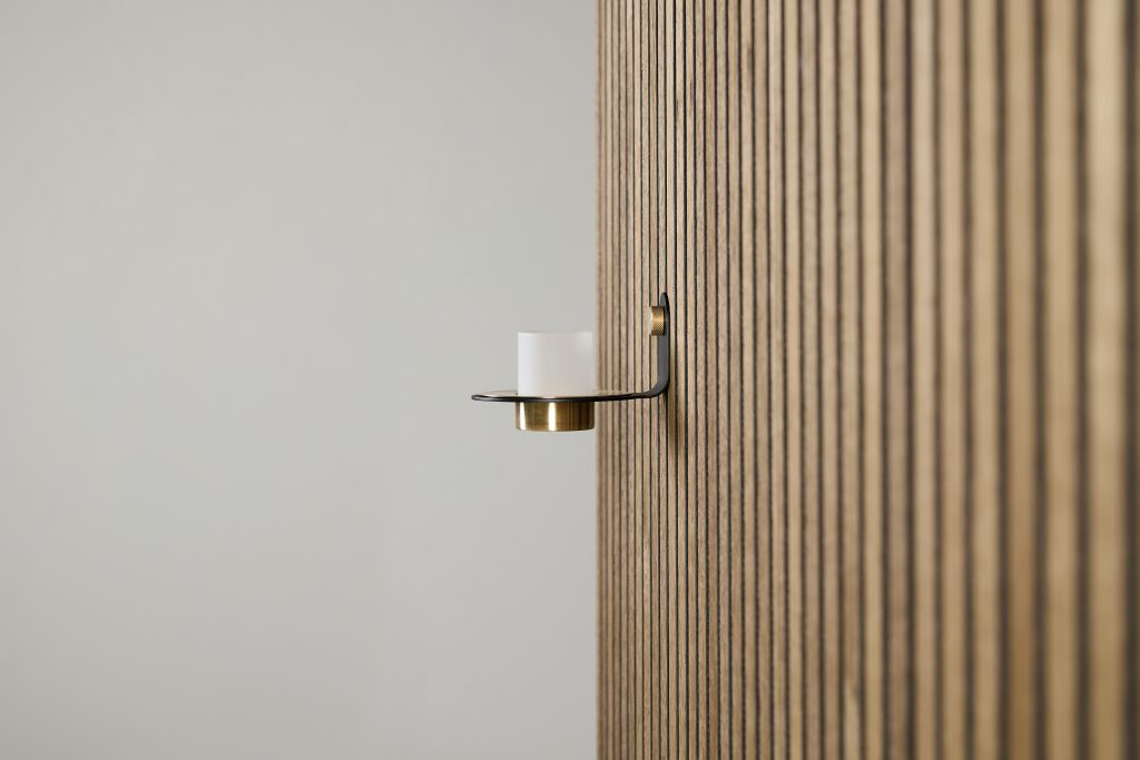 Painel ripado em madeira com suporte de vela WoodUpp: arandela minimalista em metal preto com detalhe em latão e copo de vidro fosco para vela. Produto WoodUpp fixado na parede, combinando design elegante e acabamento quente junto às lâminas verticais de madeira.