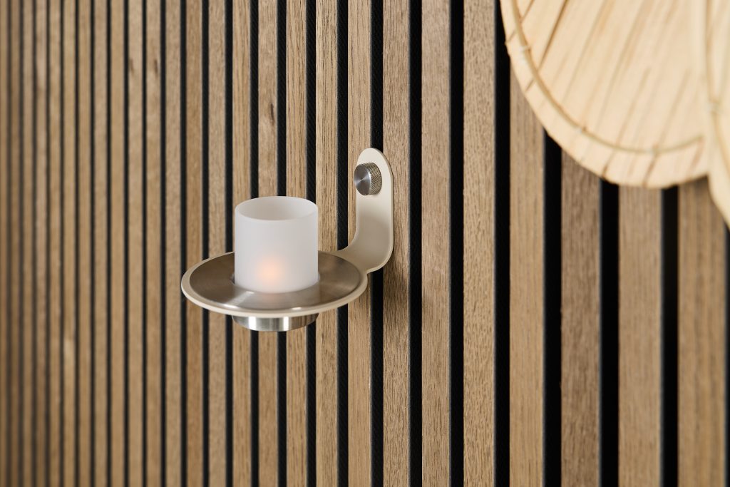 Painel ripado WoodUpp em madeira clara com ranhuras verticais e fundo preto, instalado numa parede. Ao centro, suporte de vela metálico WoodUpp com prato circular e copo de vidro fosco iluminado por uma vela LED, compondo acabamento moderno e aconchegante.