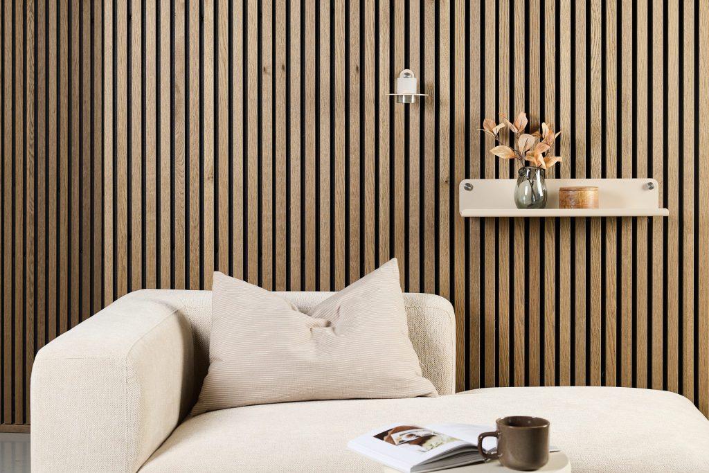 Imagem de ambiente com painel ripado de madeira WoodUpp em tons carvalho, e prateleira flutuante WoodUpp bege fixada à parede com vaso, caixa de madeira e vela; sofá creme com almofada, mesa baixa com livro aberto e caneca, em estilo minimalista e elegante.