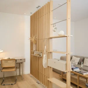 Painel ripado modular da WoodUpp em madeira clara, usado como divisória: lâminas verticais com suportes metálicos, prateleiras finas integradas, barra de metal para pendurar canecas e ganchos, e uma caixa organizadora suspensa; integrado a um escritório e sala com cadeira, escrivaninha e sofá.