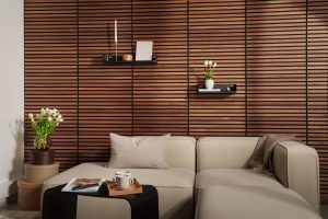 Painel ripado em madeira WoodUpp cobre a parede ao fundo; duas prateleiras flutuantes negras WoodUpp com botões metálicos exibem livros e um vaso de tulipas brancas. À frente, sofá modular bege, mesa lateral com bandeja e objetos decorativos completam a cena.