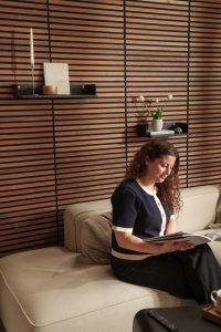 Mulher sentada em sofá bege lendo um livro diante de painel de ripas de madeira WoodUpp; duas prateleiras flutuantes pretas WoodUpp exibem vela alta, livro "Life Styled", pequeno recipiente e vaso com tulipas, com detalhes em metal dourado, compondo ambiente acolhedor e minimalista.