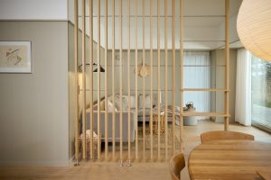 Divisória ripada WoodUpp em madeira clara, composta por lâminas verticais fixadas por hastes metálicas, separando sala e jantar; à direita, prateleiras modulares WoodUpp integradas com acabamento natural e linhas simples. Ambiente minimalista com sofá cinza, tapete rústico e iluminação suave.