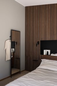 Quarto minimalista com painéis ripados WoodUpp em madeira escura na parede e cabeceira, nicho preto embutido e pequenas prateleiras WoodUpp, espelho de corpo inteiro com moldura preta, luminária de leitura e cama com roupa branca; objetos decorativos e livros sobre a cabeceira.