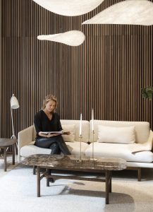 Interior WoodUpp com painel ripado de madeira escura WoodUpp cobrindo toda a parede; sofá modular bege WoodUpp com almofadas; mesa de centro de madeira escura com tampo em mármore; candelabros dourados e luminárias pendentes brancas. Mulher sentada lendo revista no sofá.