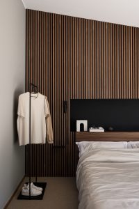 Quarto minimalista com painel ripado de madeira escura WoodUpp cobrindo a parede, cabeceira WoodUpp em madeira com nicho preto integrado, cama com roupa de cama branca e prateleira superior com livros e objetos decorativos; ao lado, cabideiro preto e um par de tênis no chão.