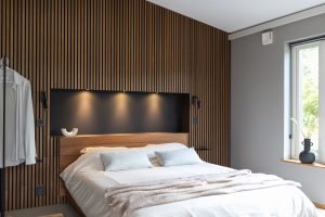 Quarto minimalista com cama branca, manta bege e travesseiros, painel ripado vertical WoodUpp em madeira escura cobrindo a parede e cabeceira WoodUpp integrada, nicho preto com três arandelas embutidas, luminárias de leitura pretas, vaso na janela e objeto decorativo branco no nicho.