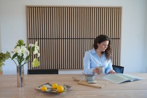 Painel ripado WoodUpp de madeira clara ocupa a parede ao fundo, com lâminas verticais finas e moldura preta, efeito acústico e decorativo. À frente, mesa de madeira clara, vaso com flores, fruteira com limões e mulher lendo, segurando uma caneca.