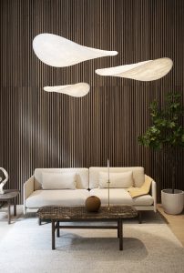 Sala contemporânea com parede ripada WoodUpp em madeira escura, três painéis acústicos suspensos WoodUpp de formato orgânico e tecido branco, sofá claro, mesa de centro em madeira escura, tapete neutro e planta em vaso. Iluminação suave destaca texturas e linhas verticais.