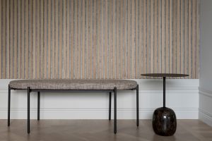 Painel acústico WoodUpp de ripas verticais em madeira clara sobre fundo cinza, cobrindo a parede; à frente, um banco estofado neutro com estrutura metálica preta e uma mesa lateral com base de pedra escura e tampo fino — ambiente minimalista e elegante.