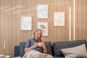 Interior com parede ripada WoodUpp: painéis verticais de madeira clara WoodUpp cobrindo toda a parede, acompanhados por quadros decorativos de traço minimalista em moldura de madeira; sofá cinza com almofadas e manta, mulher sentada segurando uma caneca, e iluminação linear embutida à direita.