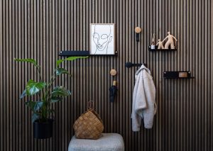 Painel de ripas de madeira com produtos WoodUpp: prateleiras pretas modulares fixadas com botões metálicos, cabideiros combinando haste preta e botão de madeira (um com suporte esférico para casaco, outro sustentando guarda-chuva), e um suporte pendente para vaso com planta, criando entrada funcional e decorativa.