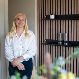 Mulher loira sorridente em pé junto a um painel ripado de madeira WoodUpp, com duas prateleiras metálicas pretas WoodUpp fixadas por botões dourados; nas prateleiras há uma vela/caixinha e dois copos de vidro estriado, em ambiente claro e acolhedor moderno.