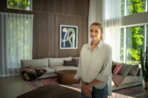 Mulher em sala de estar moderna, com painel ripado de madeira WoodUpp atrás do sofá, mesa de centro redonda em madeira WoodUpp, sofá neutro com almofadas, cortinas claras e amplas janelas que revelam jardim. Ambiente luminoso e acolhedor, com tapete e plantas.