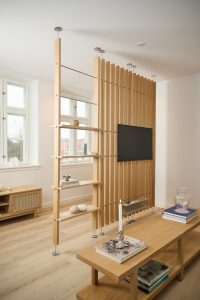 Divisória WoodUpp em ripas de madeira clara, montada entre teto e piso com hastes metálicas, com painéis verticais e prateleiras integradas; suporte para TV embutido nas ripas. Ambiente de sala de estar com móveis em carvalho claro, objetos decorativos e iluminação natural.