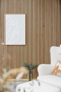 Interior minimalista com painel ripado de madeira claro da WoodUpp, com ripas verticais e cavidades pretas entre elas, moldura de quadro em madeira natural e rodapé combinando; quadro branco de textura ondulada, vaso de vidro com folhagens e poltrona bouclé com almofada estampada.