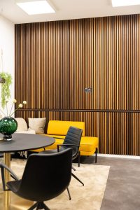 Interior com painéis ripados WoodUpp cobrindo toda a parede: ripas verticais em madeira tom carvalho com frestas escuras, criando efeito acústico e estético. Ambiente com sofá amarelo mostarda, cadeiras pretas e mesa redonda, destacando o acabamento moderno dos produtos WoodUpp.