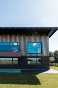 Fachada moderna com revestimento ripado vertical WoodUpp no piso superior, grandes janelas com caixilhos escuros e forro de beiral em painéis de madeira WoodUpp; sacada com guarda-corpo de vidro e alvenaria de tijolo escuro no pavimento térreo, em frente a gramado e céu claro.