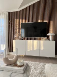 Interior minimalista com painel ripado WoodUpp em ripas de madeira escura vertical cobrindo a parede atrás de uma TV de tela plana; móvel suspenso branco, vaso de vidro com galho seco, luminária branca e objetos decorativos sobre o aparador, tapete felpudo e vela em mesa baixa.