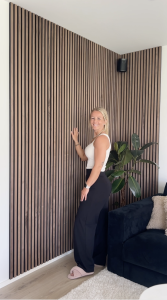Uma mulher sorridente está encostada a uma instalação de canto revestida com painéis acústicos da WoodUpp. Os painéis apresentam ripas de madeira escura, estilo nogueira, sobre um fundo de feltro preto, cobrindo as paredes do chão ao teto. Uma planta alta e um sofá de veludo azul completam o ambiente moderno.