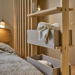 Divisória estante WoodUpp em madeira clara com prateleiras finas e caixas organizadoras de feltro cinza encaixadas, mostrando um suéter creme dobrado e espaço inferior com roupa; ao lado, cama acolchoada e luminária de cabeceira completam o ambiente acolhedor do quarto.