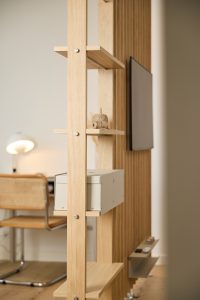 Painel divisório WoodUpp em madeira clara com prateleiras finas e parafusos aparentes, suporte para TV e gaveta metálica modular WoodUpp cinza instalada; na prateleira há um novelo de barbante. Ao fundo vê-se uma secretária com cadeira de rattan e abajur.