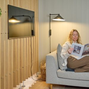 Interior aconchegante com painéis ripados WoodUpp em madeira clara cobrindo a parede e servindo de fundo para a TV; estrutura de suporte metálica na base é visível. À direita, sofá cinza com mulher lendo revista sob luminária, cesto de vime no chão.