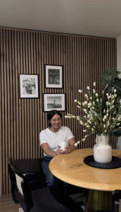 Uma mulher sorri com uma caneca numa mesa redonda de madeira. Atrás, uma parede revestida com os painéis acústicos ripados de madeira da WoodUpp, em tom de nogueira com fundo de feltro preto, decorada com três quadros e um vaso de flores brancas.