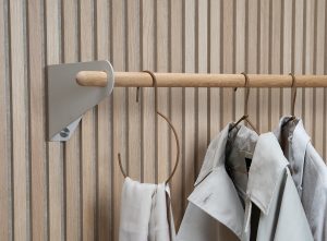 Imagem WoodUpp mostrando painéis ripados de madeira clara, um varão de cabide em carvalho e o suporte de parede metálico triangular WoodUpp; sobre o varão, cabides em couro minimalistas e casacos em tons neutros, numa composição clean, sofisticada e elegante.