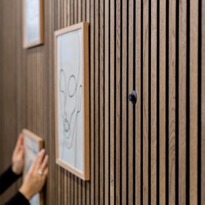 Painel ripado de madeira WoodUpp instalado na parede, com nervuras verticais regulares. Em destaque, um pequeno gancho/pendurador preto da WoodUpp preso entre as ripas, enquanto mãos penduram quadros em moldura de madeira à esquerda, mostrando sistema de decoração e organização WoodUpp.